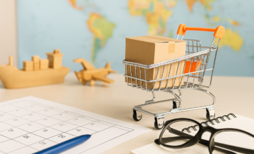 Carrinho de compras com caixa de papelão, miniaturas de navio e avião, calendário, caderno e óculos sobre mesa, representando planejamento de importação e logística internacional.