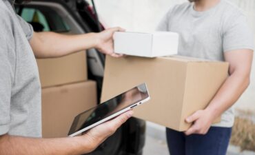 Entregador realizando entrega com caixas enquanto outro profissional confirma dados em um tablet.